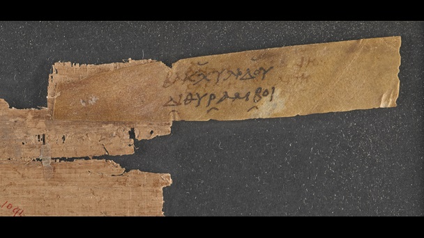 A small fragment of a 2nd-century papyrus scroll, with an attached parchment sillybos, or label, inscribed with the title of a work by Bacchylides.
