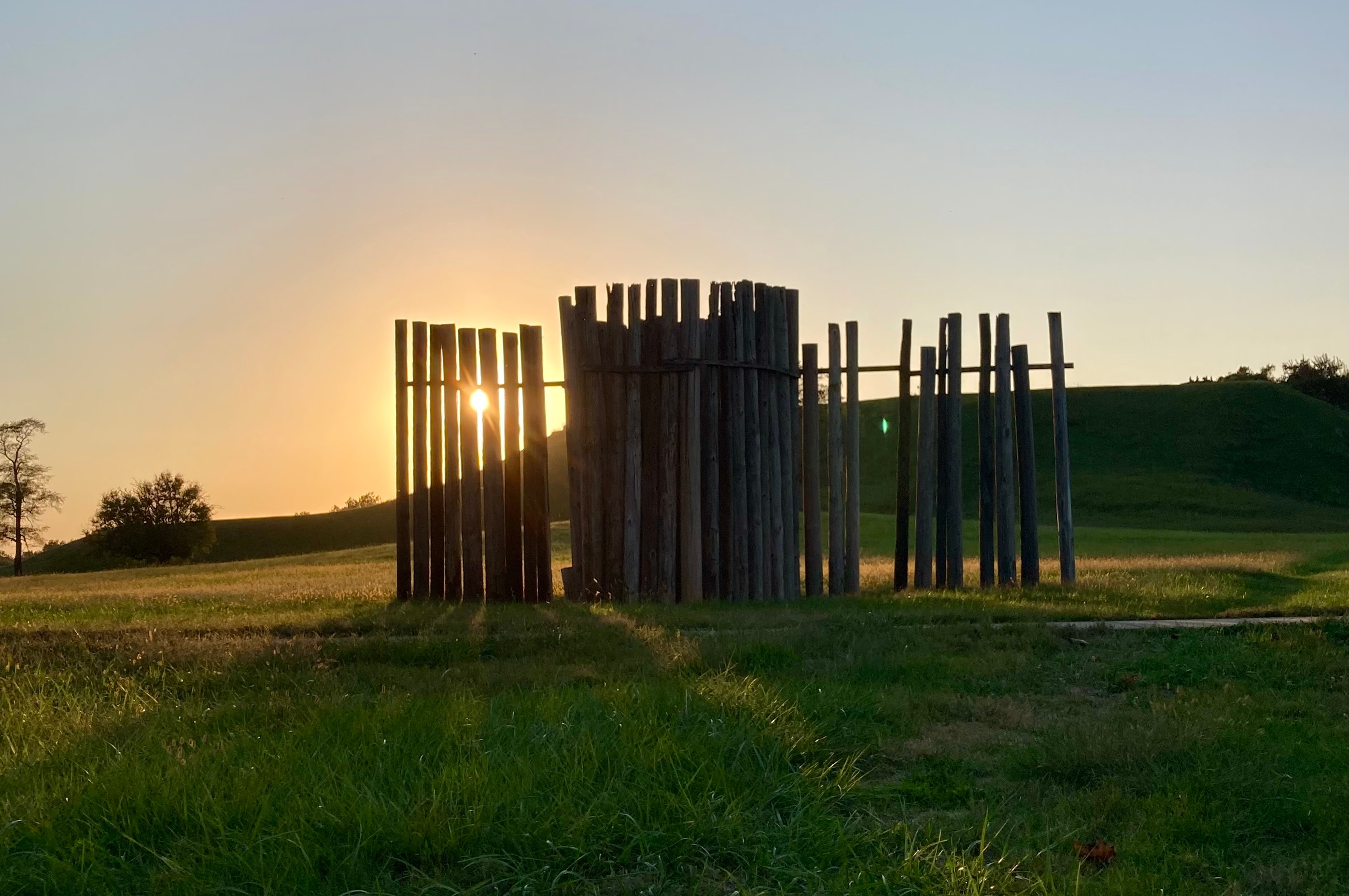 An orange sunset illuminates Monks Mound through a stand of wooden posts.